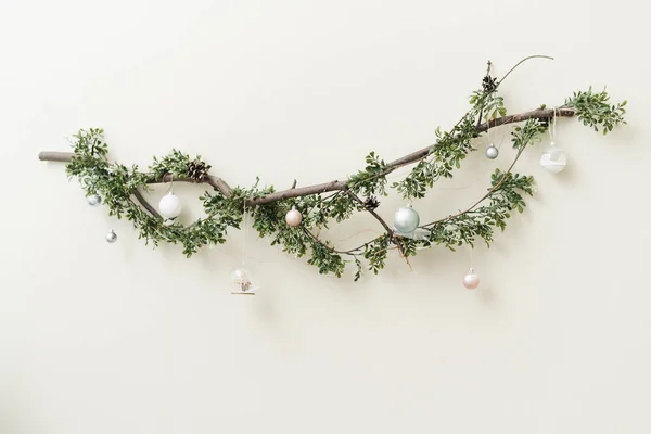 Ein dünner Ast mit einer grünen Weihnachtsgirlande, silbernen und goldenen Weihnachtskugeln und Spielzeug. schöne Weihnachtsdekoration an einer weißen Wand — Stockfoto