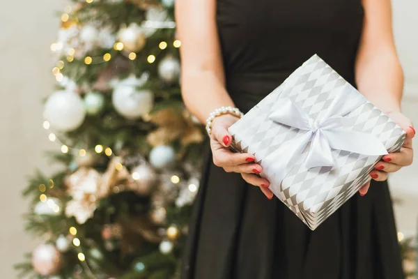 Uma menina em um vestido preto festivo no fundo de uma árvore de Natal com bokeh detém um presente e dá-lo para a câmera. Uma mulher dá um presente de Ano Novo — Fotografia de Stock