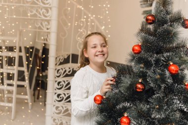Şirin küçük kız Noel ağacını yeni yıl oyuncakları ve kırmızı toplarla süslüyor. Beyaz örgü süveter ve elbise giyen bir kız yapay bir ladin üzerinde asılı duran toplar.