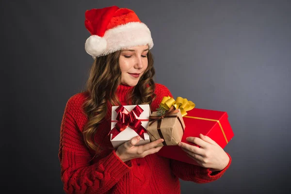 Una hermosa modelo en un sombrero de Santa Claus y un suéter de punto rojo tiene muchos regalos en sus manos. El concepto de Año Nuevo, regalos de Navidad, dar, compras de Navidad. Plantilla para tu publicidad —  Fotos de Stock