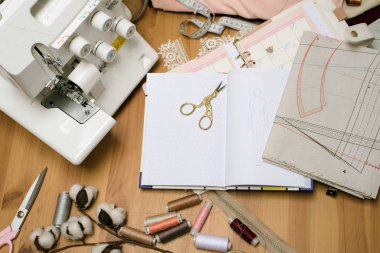 Dağılmış makaslı ahşap bir terzi masası, bir üst kilit, bir dikiş makinesi, çizimler, iplikler ve kumaşlı taslaklar. Çalışan terzi eşyalarıyla dolu bir yer.