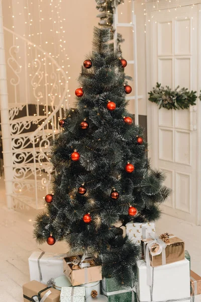 Kunstmatige kerstboom met rode ballen en decor erop. Kerst- en nieuwjaarsgeschenken onder de kerstboom in een helder interieur — Stockfoto