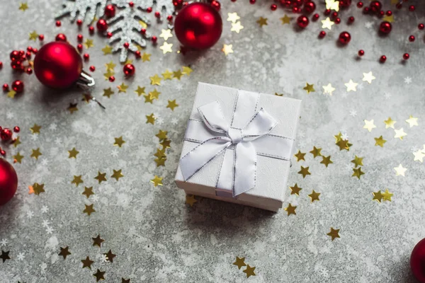 Silbergeschenk mit Schleife auf grauem, schneebedecktem Hintergrund mit roten Weihnachtskugeln und goldenen Konfetti-Sternen — Stockfoto