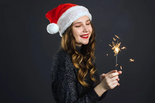 Linda chica en el sombrero de Santa se alegra de Navidad y Año Nuevo sobre un fondo gris oscuro. La chica tiene un bengala —  Fotos de Stock