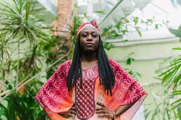 Uma jovem mulher afro-americana bonita está sob palmeiras na selva e sorri amplamente em um traje africano nacional. Retrato ao ar livre sensível da menina africana atraente com sombras — Fotografia de Stock