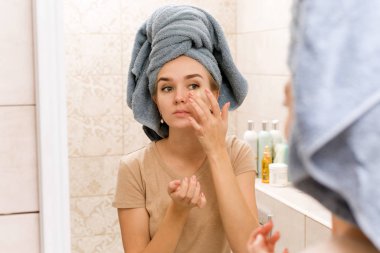 Kafasında havluyla güzel bir kız gözlerinin altına hidrojel bantlar yapıştırıyor. Kız cildi önemsiyor ve göz çevresindeki bölgeyi nemlendiriyor. Evde bakım giderleri banyoda.