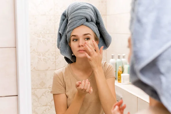 Una hermosa chica con una toalla en la cabeza le pone parches de hidrogel debajo de los ojos en la cara. La niña cuida la piel e hidrata el área alrededor de los ojos. Tratamientos de atención domiciliaria en el baño — Foto de Stock