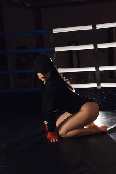 Sexy fighter girl in a black bodysuit and cap posing in a dark black boxing ring. Long hair woman fitness model. Attractive female boxer training. The concept of a healthy lifestyle