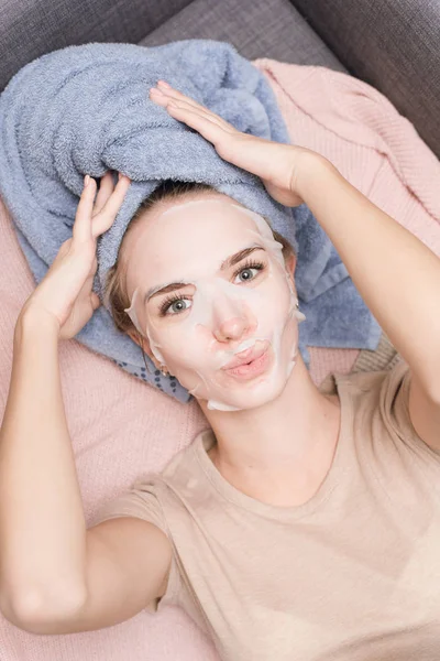 Linda chica encantadora está acostada en el sofá en un tejido desechable máscara hidratante y toalla en la cabeza y sonriendo. Cuidado en el hogar, cuidado de la piel y tratamientos antienvejecimiento — Foto de Stock