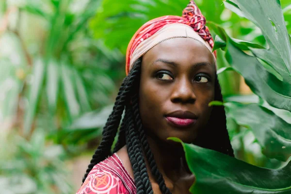 Egy csinos fiatal afro-amerikai nő áll a pálmafák alatt a dzsungelben, és széles körben mosolyog egy nemzeti afrikai jelmezben. Érzékeny kültéri portré a vonzó afrikai lányról szemhéjpúderekkel — Stock Fotó