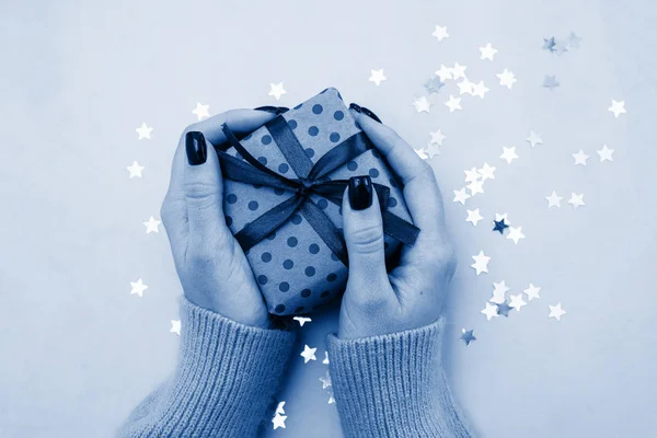 Classic blue, toned image. A woman hands holding present, manicured hands with nail polish. Girl gives a gift on a blue background with confetti. Trend of the season 2020. Classic blue is the New colo