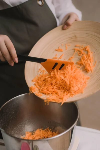 Повар добавляет тертую морковь в металлическую миску. Женская рука кладет нарезанную морковку в деревянную миску с салатом на кухне. Кулинария — стоковое фото