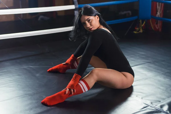 Sexig fighter flicka i svart body suit och röd boxning bandage och leggings poserar i en mörk svart boxning ring. Lång hår kvinna fitness modell. Begreppet en hälsosam livsstil. — Stockfoto