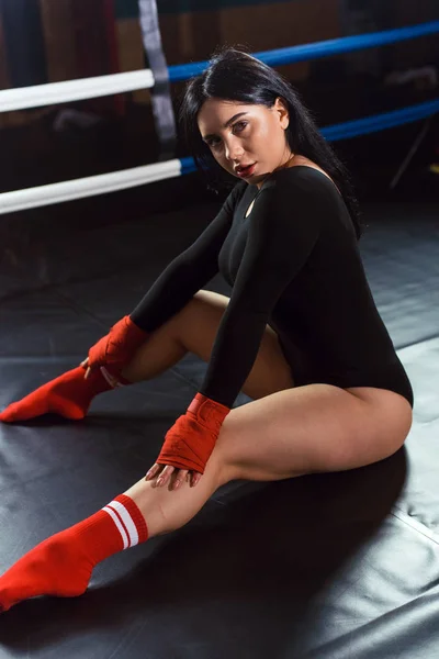 黒のボディスーツと赤のボクシングの包帯とレギンスでセクシーな戦闘機の女の子は暗い黒のボクシングリングにポーズ。ロングヘア女性フィットネスモデル。健康的なライフスタイルの概念 — ストック写真
