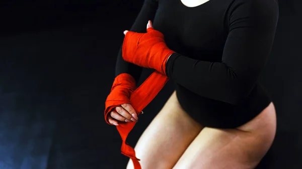 Sexy woman is wrapping hands with red boxing wraps. Strong hand and fist, ready for fight and active exercise. Women self defense. A girl in a black body wraps hands with bandages. The concept of femi — Stock Photo, Image