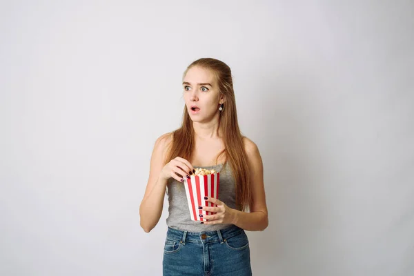 La chica se sorprende y mira a la cámara, comiendo palomitas de maíz sobre un fondo gris blanco. Plantilla para un blog sobre películas o anuncios de cine —  Fotos de Stock