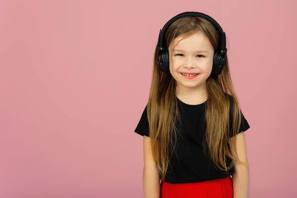 Una niña pequeña con auriculares negros grandes inalámbricos sobre un fondo rosa pastel mira a la cámara. El concepto de escuchar y disfrutar de la música. Lugar para texto, copyspace — Foto de Stock