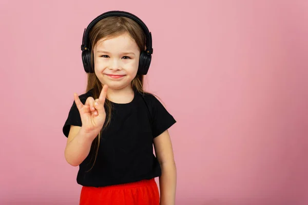 Una niña pequeña con auriculares negros grandes inalámbricos sobre un fondo rosa pastel mira a la cámara y muestra un gesto de cuernos hacia arriba. El concepto de escuchar música rock. Lugar para texto, copyspace — Foto de Stock