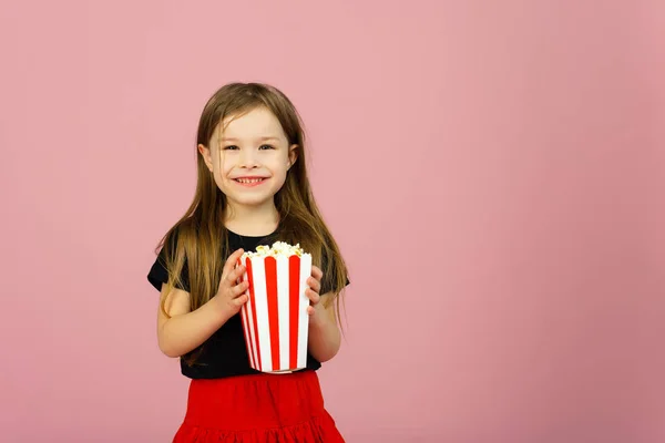 Ευτυχισμένο χαριτωμένο κοριτσάκι που κρατάει ποπ κορν στα χέρια της και κοιτάζει την κάμερα σε ροζ φόντο. Η ιδέα του να βλέπεις μια ταινία σε κινηματογράφο ή στο σπίτι. Αντιγραφικός χώρος — Φωτογραφία Αρχείου