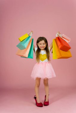 Güzel, sarı saçlı, uzun etekli, annesinin yanında duran sevimli küçük bir kız. Pembe arka planda izole edilmiş renkli paketleri olan büyük ayakkabılar. Kameraya gerçek olumlu duyguları ifade etmek.