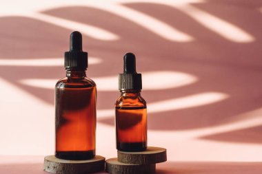 Tahta zeminde nemlendirici serumu olan iki cam şişe pembe arka planda kesilmiş. Palmiye yaprağının tropikal gölgesinde kahverengi tüpler. Cilt bakımı kavramı.