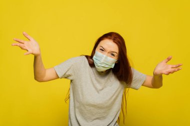 A woman clings to her head and experiences fear, shock from the spread of the disease. Red-haired beautiful girl in a protective mask on a yellow background screams from a coronavirus. The concept of protection and prevention. clipart
