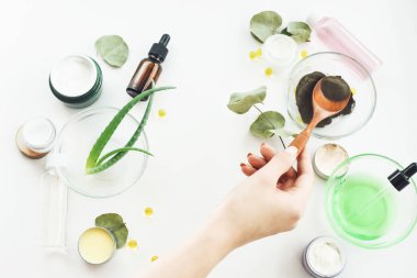 Ev yapımı ilaç ve güzellik ürünü konsepti. Aloe vera yaprakları, siyah kil tozu, C vitamini, çiçek özü, beyaz arka planda serumlu şişeler, tonikler, krema kavanozları ve jel. Kızın eli krem yapmak için malzemeleri karıştırıyor..