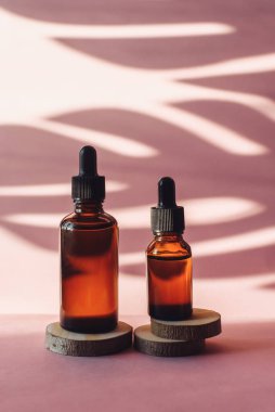Tahta zeminde nemlendirici serumu olan iki cam şişe pembe arka planda kesilmiş. Palmiye yaprağının tropikal gölgesinde kahverengi tüpler. Cilt bakımı kavramı.