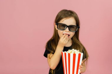 Sevimli küçük kız çizgili ambalajdan patlamış mısır yiyor ve 3D gözlüklü bir film izliyor. Sinema reklamcılığı konsepti, çevrimiçi film izlemek.