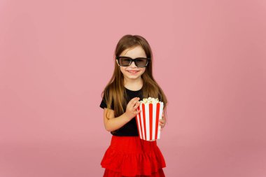 Sevimli küçük kız çizgili ambalajdan patlamış mısır yiyor ve 3D gözlüklü bir film izliyor. Sinema reklamcılığı konsepti, çevrimiçi film izlemek.