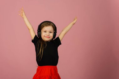 Küçük bir kız pembe pastel arka planda büyük kulaklıklarla müzik dinliyor ve ellerini kaldırarak seviniyor. Samimi duygular.