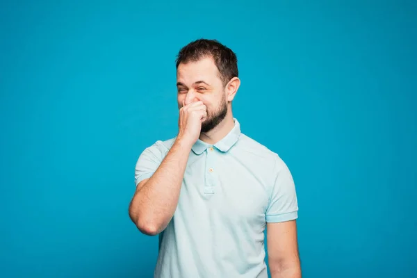 A man in a blue T-shirt on a blue background funny sneezes in his hand. You can not sneeze in the hand, you need to sneeze on the elbow or in a scarf. Violation of the rules. People, disease concept — Stock Photo, Image