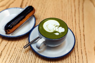 Kibrit yeşili, beyaz bardakta, bir kahve dükkanındaki ahşap masada, tatlı bir tatlının yanında. Kahvaltıda çikolatalı ekler..