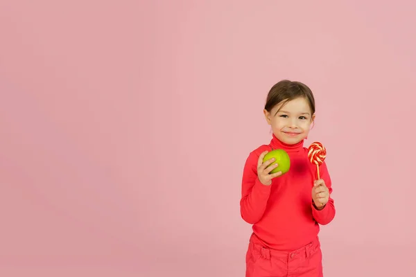Το κοριτσάκι διαλέγει ανάμεσα σε ένα γλειφιτζούρι και ένα πράσινο μήλο. Η έννοια της σωστής διατροφής. Ένα παιδί σε ένα ροζ στούντιο κρατάει μια γλυκιά ζάχαρη στο χέρι του και ένα μήλο. Δυσκολία επιλογής — Φωτογραφία Αρχείου