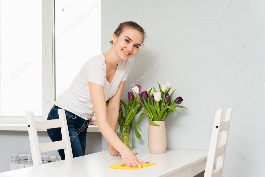 A housewife in a shirt is cleaning the house, wipes dust from the table with a cleaning rag. A cute blonde is smiling at the camera while cleaning her house.