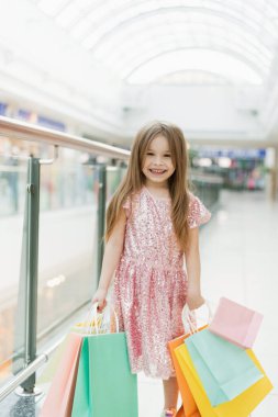 Alışveriş merkezinde küçük mutlu bir kızın portresi. Elinde çok renkli poşetler olan pembe elbiseli gülen bir kız alışverişle meşgul. Reklamınız için şablon.