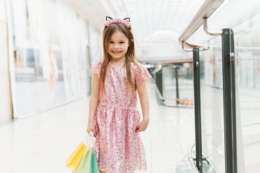 Alışveriş merkezinde küçük mutlu bir kızın portresi. Pembe elbiseli, kulakları kulakları ve ellerinde renkli çantalarıyla gülümseyen bir kız alışveriş merkezinde kameraya bakıyor..