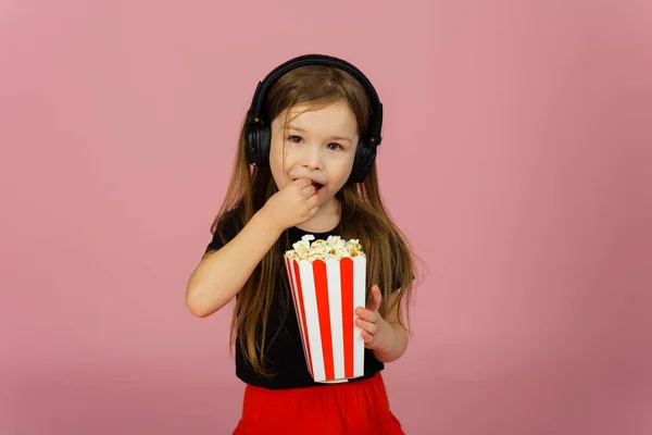 Little girl on a pink pastel background in big headphones eats pop corn in striped packaging. The concept of going to the cinema, advertising a movie theater.