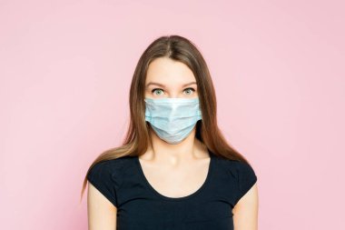Coronavirus, maskeli kadın. Stüdyo portresi, yüz maskesi takan, pembe arka planda kameraya bakan genç bir kadın..