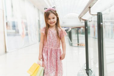 Alışveriş merkezinde küçük mutlu bir kızın portresi. Pembe elbiseli, kulakları kulakları ve ellerinde renkli çantalarıyla gülümseyen bir kız alışveriş merkezinde kameraya bakıyor..