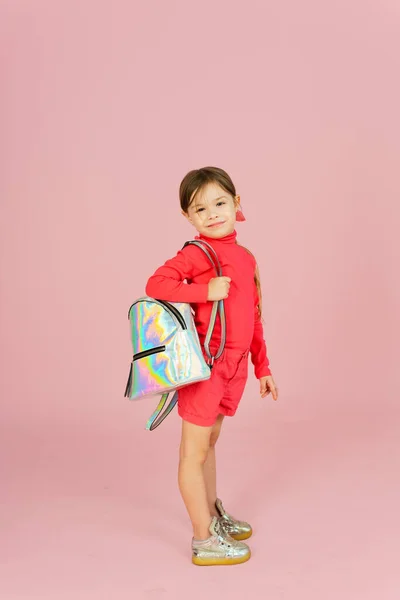 stock image Cute little blonde classmate in a pink suit in the studio with a holographic backpack. Funny first grader on a pink pastel background with copyspace.