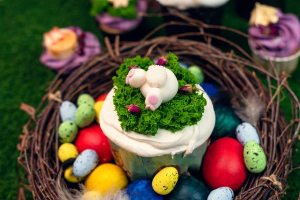 Orthodox Easter cake in a wooden nest with painted chicken and quail eggs on green grass. Easter cake with a figurine of a ponytail of a rabbit on the background of sweets, cupcakes.