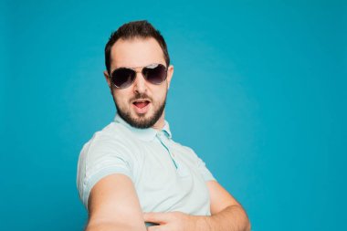 Genç, sakallı bir adam mavi arka planda kameraya bakıyor. Adam bir selfie çekip komik suratlar yapıyor..