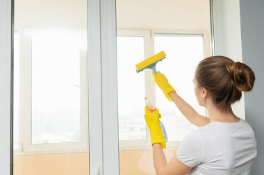 Sevimli yetişkin kız bir kazıyıcıyla pencereyi yıkıyor. Bahar temizliği. Temizlik şirketinin bir çalışanı evdeki plastik camları siliyor..