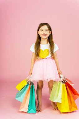 Pastel pembe arka planda bir sürü alışveriş torbası olan kız. Küçük prensesin sevimli tatlı anları, oldukça arkadaş canlısı çocuk kamerayla eğleniyor..
