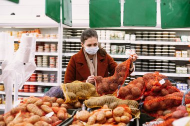 Pandemik karantina hazırlığı. Kuru ürünler ve besin maddeleri alışverişi. Koruyucu maskeli genç kadın patates ve sebze seçiyor, süpermarketten ürünler alıyor, hisse senetleri karantinaya alınıyor..