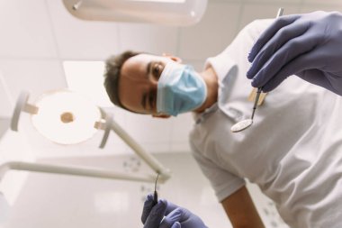 Profesyonel diş hekimi ofisinde çalışıyor. Koruma eldivenleri ve maskeli genç bir dişçi, dişçi aletini kameraya bakmaya devam ediyor..