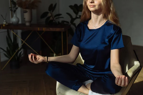 Young woman meditating in armchair at home. Young women meditating with mudra sign at yoga class or her home. Girl practice sitting on armchair with lotos pose crossed legs