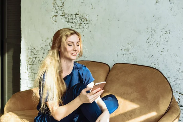 A young woman surfs the internet on the phone and smiles and laughs. Browse social networks at home while sitting on the couch