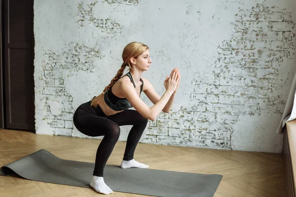 Cute blonde is engaged in fitness on the background of a white brick wall at home during quarantine. Stay home and exercise your body — Stock Photo, Image